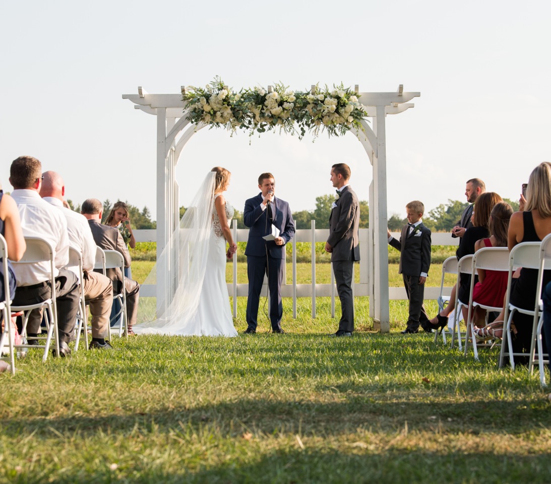 An outdoor ceremony beneath a white floral arbor with guests seated on the lawn
